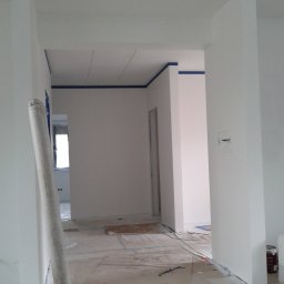 Ogólno-Budowlana - Wnętrze w trakcie odświeżania: świeżo pomalowane na biało ściany z niebieską taśmą malarską przy suficie, zabezpieczona podłoga, widoczna rolka do malowania oparta o ścianę, butelka po wodzie...