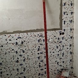 Ogólno-Budowlana - Ściana łazienki w trakcie wykańczania, z ułożonymi płytkami o wzorze lastryko, poziomica oparta o ścianę, widoczne niebieskie klipsy do poziomowania płytek, poniżej dwa wiadra i zwinięty kabel...