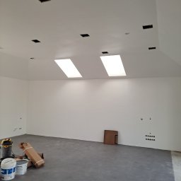 Ogólno-Budowlana - Wnętrze w trakcie prac wykończeniowych: surowy beton na podłodze, białe ściany z widocznymi puszkami elektrycznymi, otwory w suficie przygotowane pod oświetlenie, dwa świetliki, farby i odkurzacz...