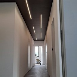 Ogólno-Budowlana - Długi korytarz z szarym sufitem, w którym zamontowano liniowe, ledowe oświetlenie. Ściany białe, podłoga betonowa. W tle widoczny odkurzacz.