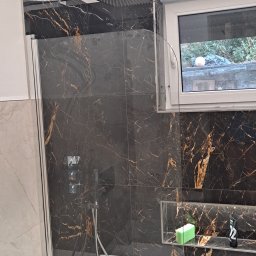 Ogólno-Budowlana - Nowoczesna łazienka z czarnymi płytkami imitującymi marmur z złotymi żyłami, kabina prysznicowa ze szklaną ścianką, deszczownica, wnęka z kosmetykami i oświetleniem LED.