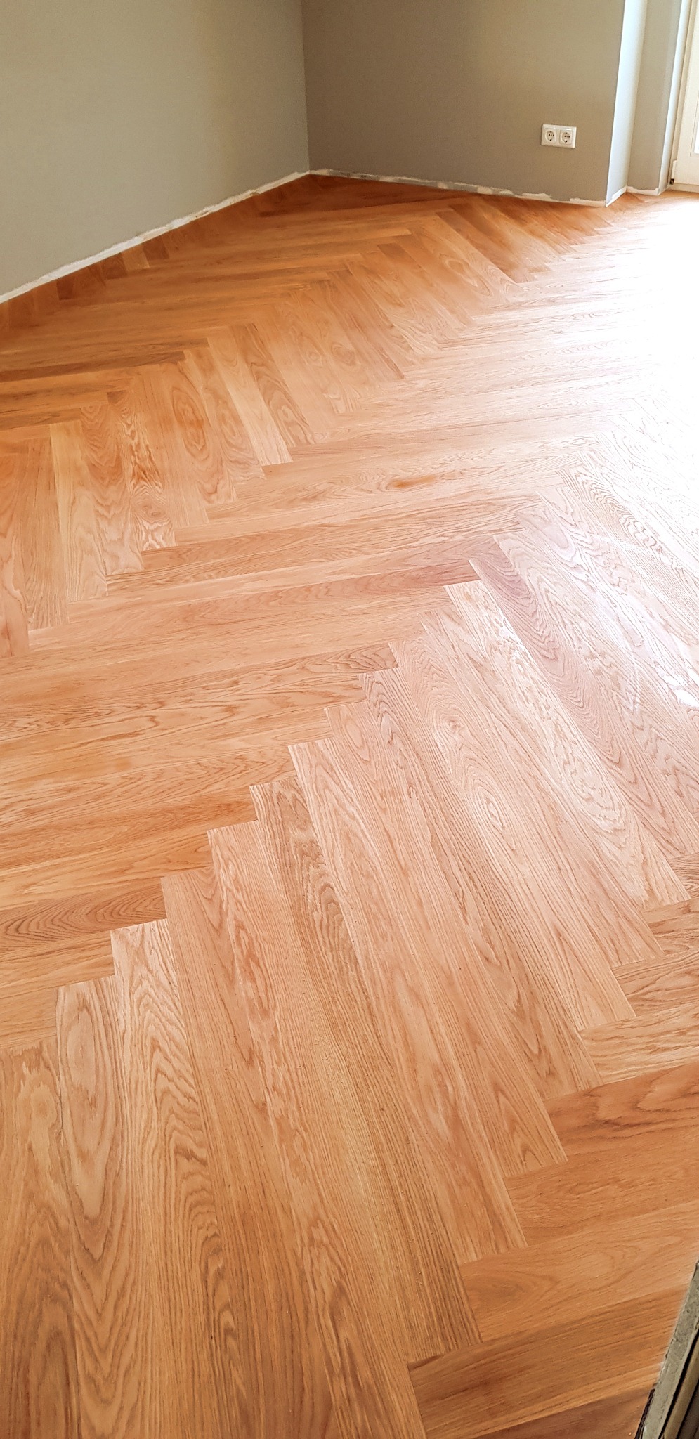 Ułożony parkiet w jodełkę francuską, jasne drewno, widoczne gniazdko elektryczne na szarej ścianie, świeżo pomalowane listwy przypodłogowe.