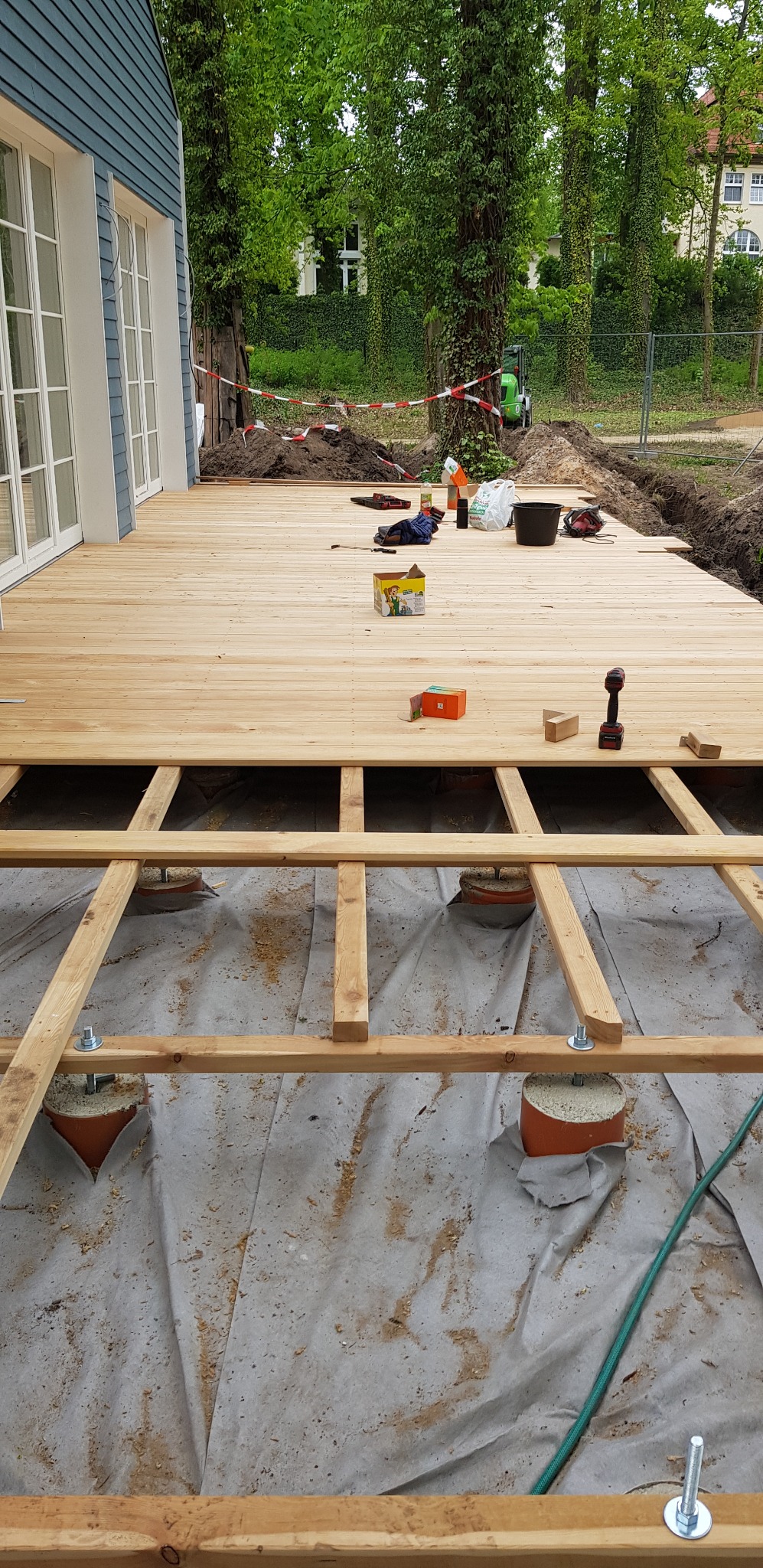 Montaż drewnianego tarasu: widok na częściowo ułożone deski na legarach, z narzędziami i materiałami budowlanymi na powierzchni, w tle trawnik i drzewa.