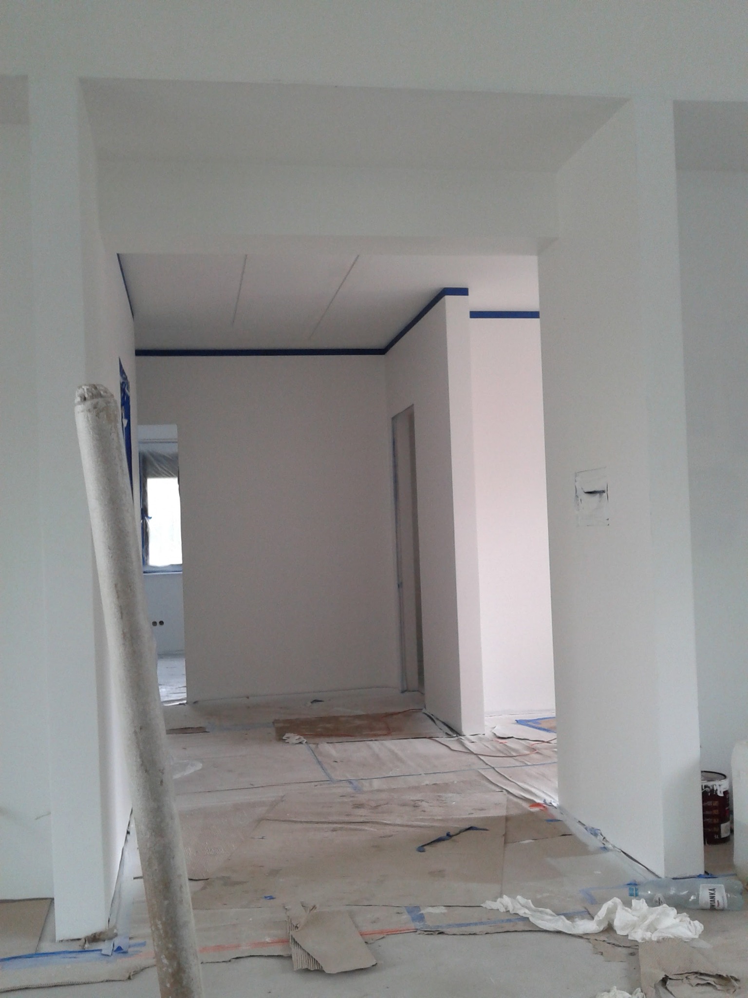 Wnętrze w trakcie odświeżania: świeżo pomalowane na biało ściany z niebieską taśmą malarską przy suficie, zabezpieczona podłoga, widoczna rolka do malowania oparta o ścianę, butelka po wodzie...