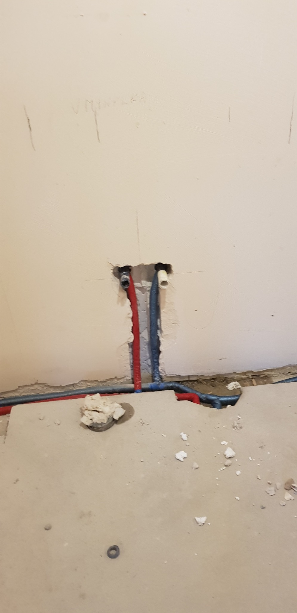 Fragment ściany w trakcie prac hydraulicznych z widocznymi przewodami w kolorach czerwonym i niebieskim, wystającymi z otworu w ścianie, oraz fragmentami gruzu na podłodze.