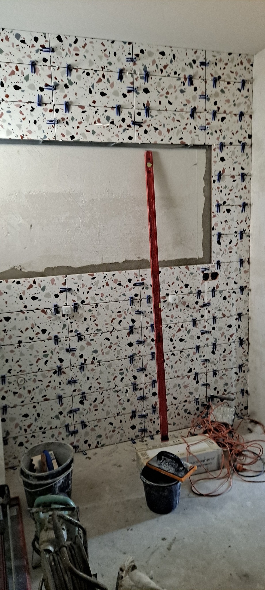 Ściana łazienki w trakcie wykańczania, z ułożonymi płytkami o wzorze lastryko, poziomica oparta o ścianę, widoczne niebieskie klipsy do poziomowania płytek, poniżej dwa wiadra i zwinięty kabel...