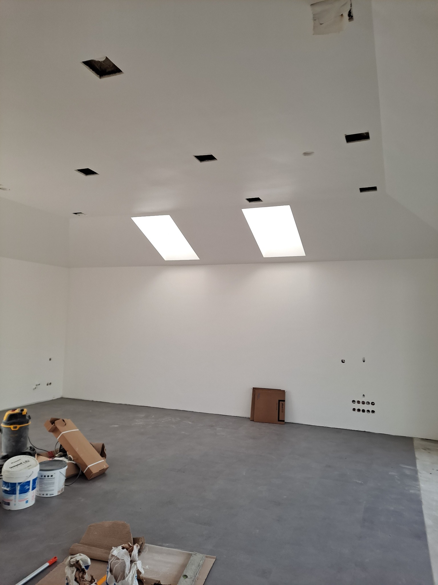 Wnętrze w trakcie prac wykończeniowych: surowy beton na podłodze, białe ściany z widocznymi puszkami elektrycznymi, otwory w suficie przygotowane pod oświetlenie, dwa świetliki, farby i odkurzacz...