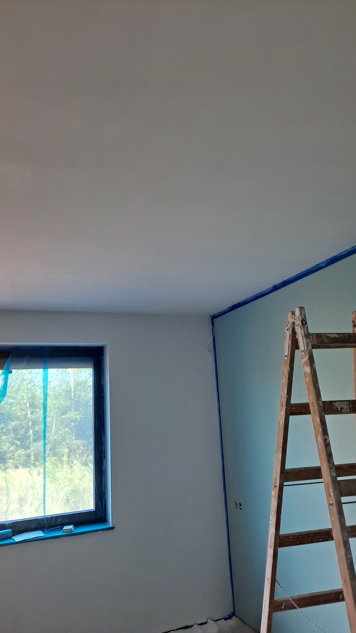 Świeżo pomalowane wnętrze pokoju z drewnianą drabiną opartą o ścianę, zabezpieczone taśmą malarską narożniki i okolice okna, widok na zieleń za oknem.