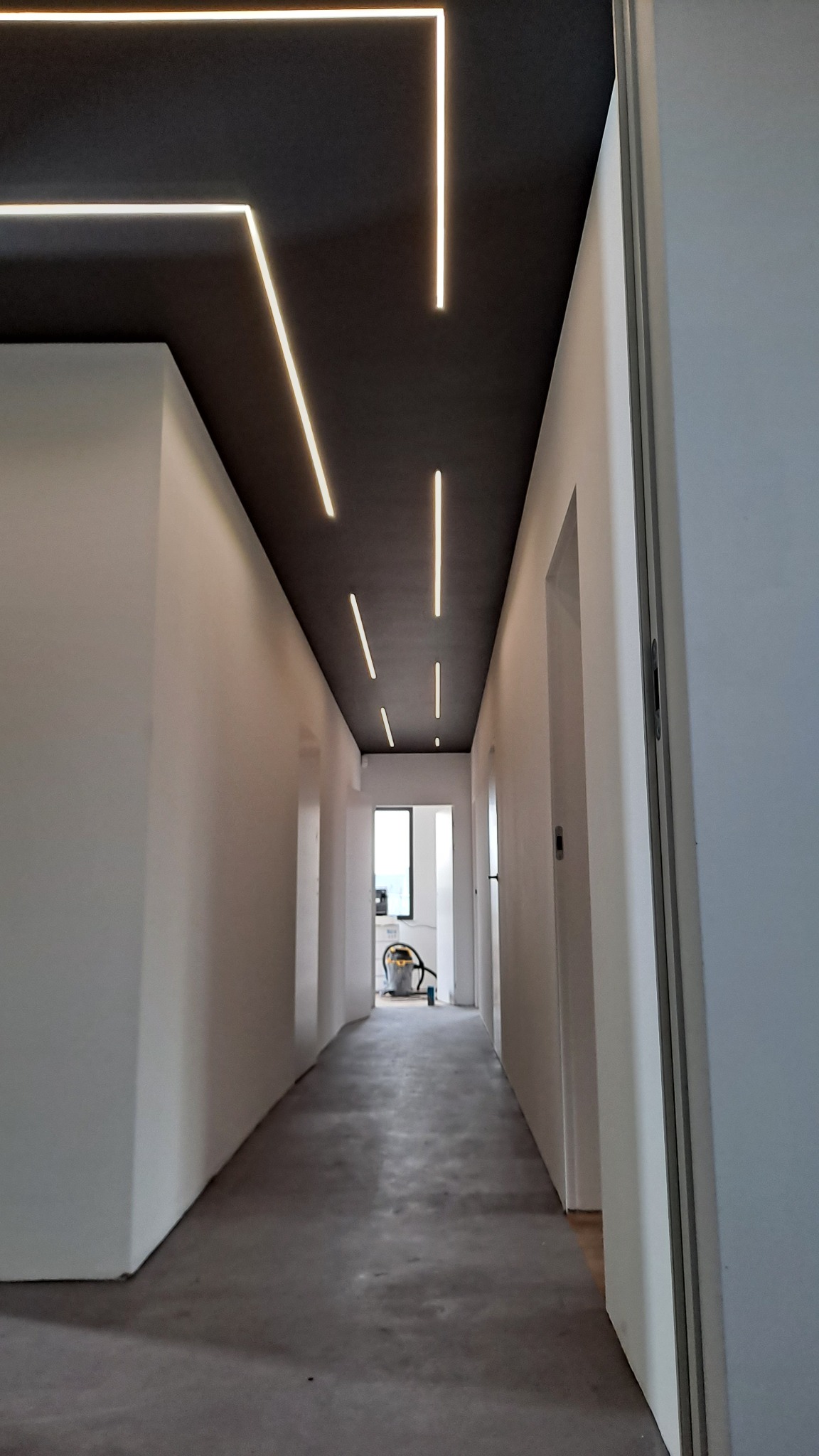 Długi korytarz z szarym sufitem, w którym zamontowano liniowe, ledowe oświetlenie. Ściany białe, podłoga betonowa. W tle widoczny odkurzacz.