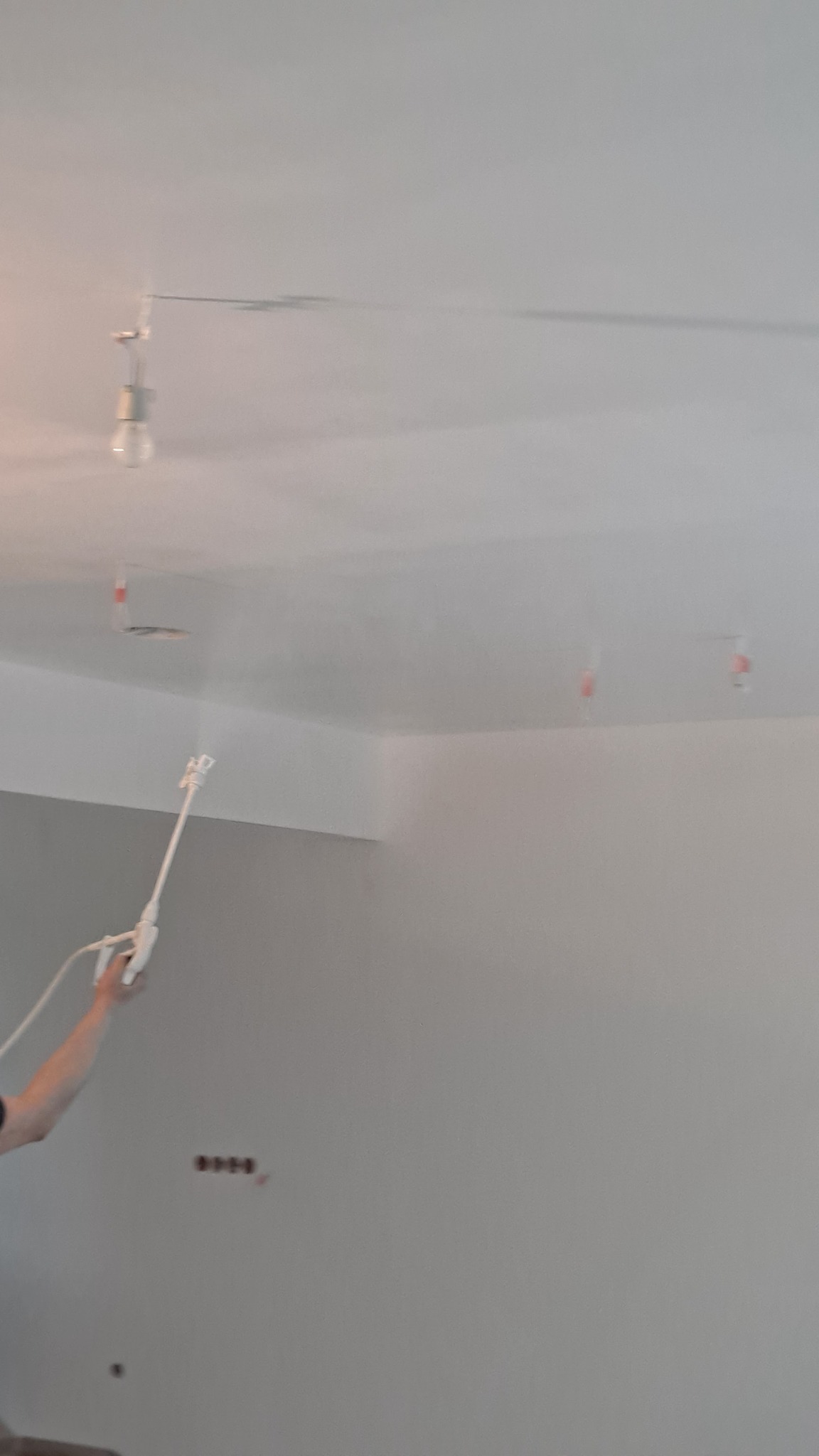 Malowanie sufitu agregatem natryskowym na biało, widoczne zabezpieczenia żarówek folią, ściana szara z gniazdkami elektrycznymi.