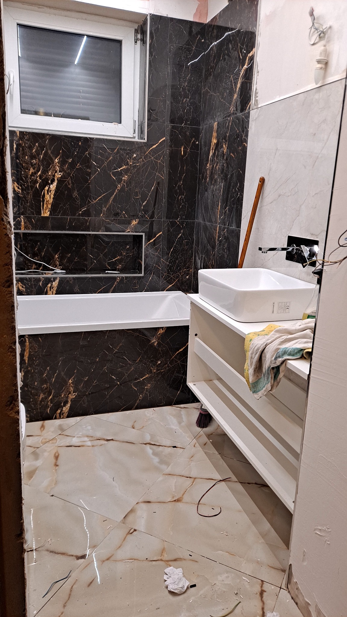 Łazienka w trakcie prac: czarne płytki imitujące marmur na ścianach, biała wanna i umywalka na szafce, podłoga w beżowych płytkach z wzorem marmuru, widoczne ślady po remoncie.