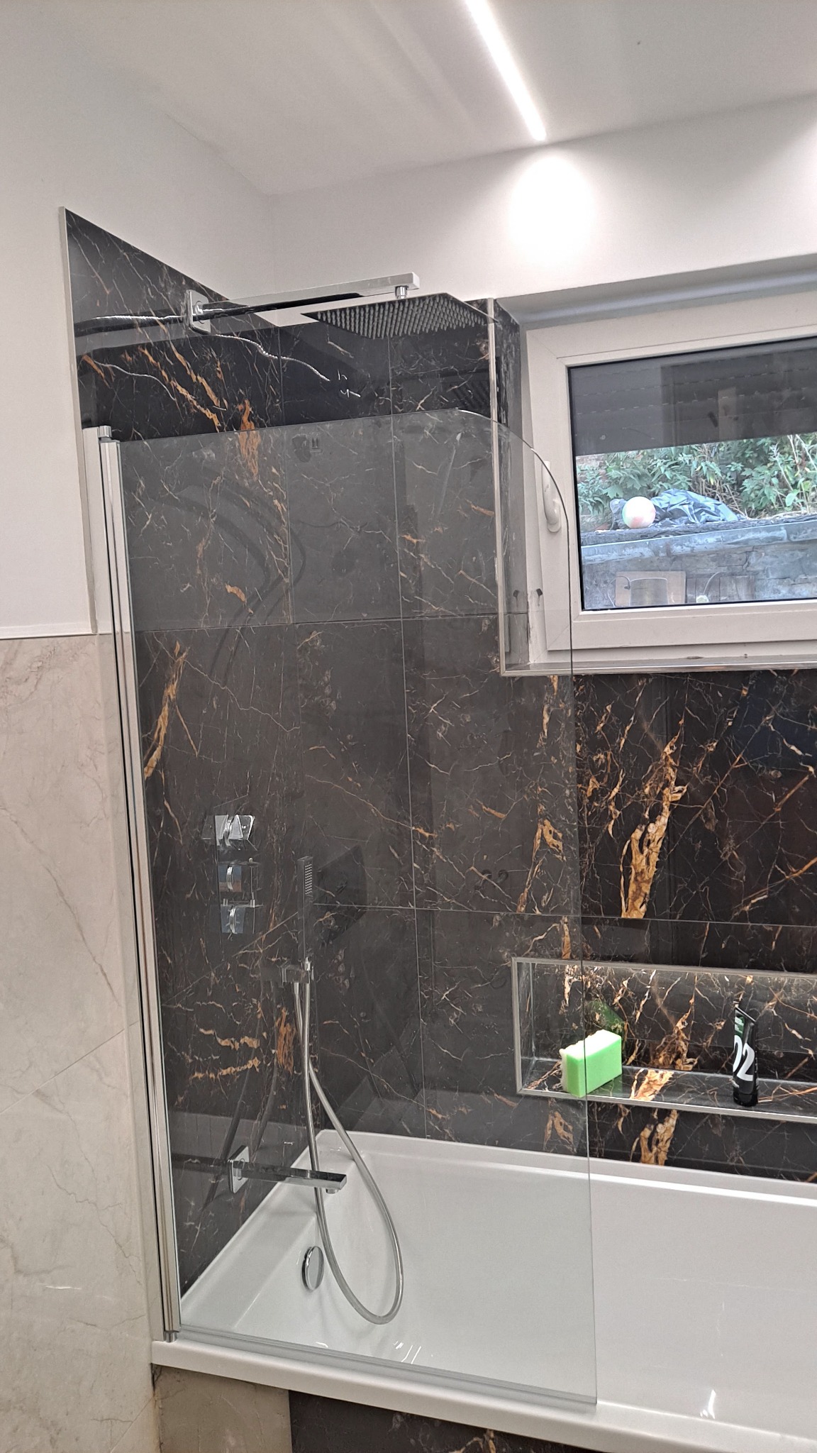 Nowoczesna łazienka z czarnymi płytkami imitującymi marmur z złotymi żyłami, kabina prysznicowa ze szklaną ścianką, deszczownica, wnęka z kosmetykami i oświetleniem LED.