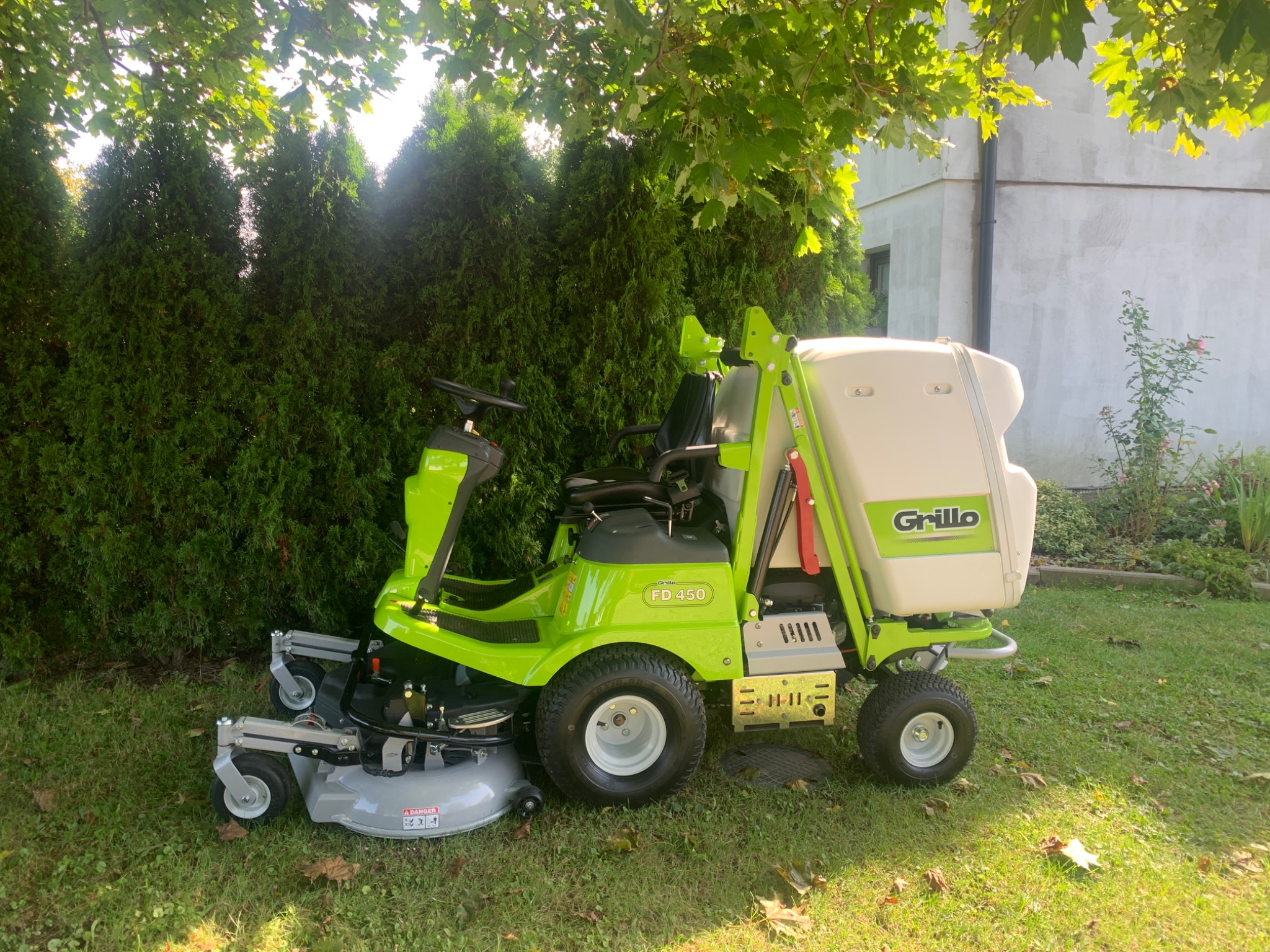Zielona kosiarka samojezdna Grillo FD 450 na trawniku przed wysokimi tujami i jasnym budynkiem, widoczny fragment trawnika z opadłymi liśćmi.