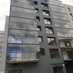 M-BUD Magdalena Lipigórska - Nowoczesny budynek wielorodzinny z szarymi panelami elewacyjnymi i przeszklonymi balkonami, w których odbija się niebo. Widok z dołu, lekki kąt.