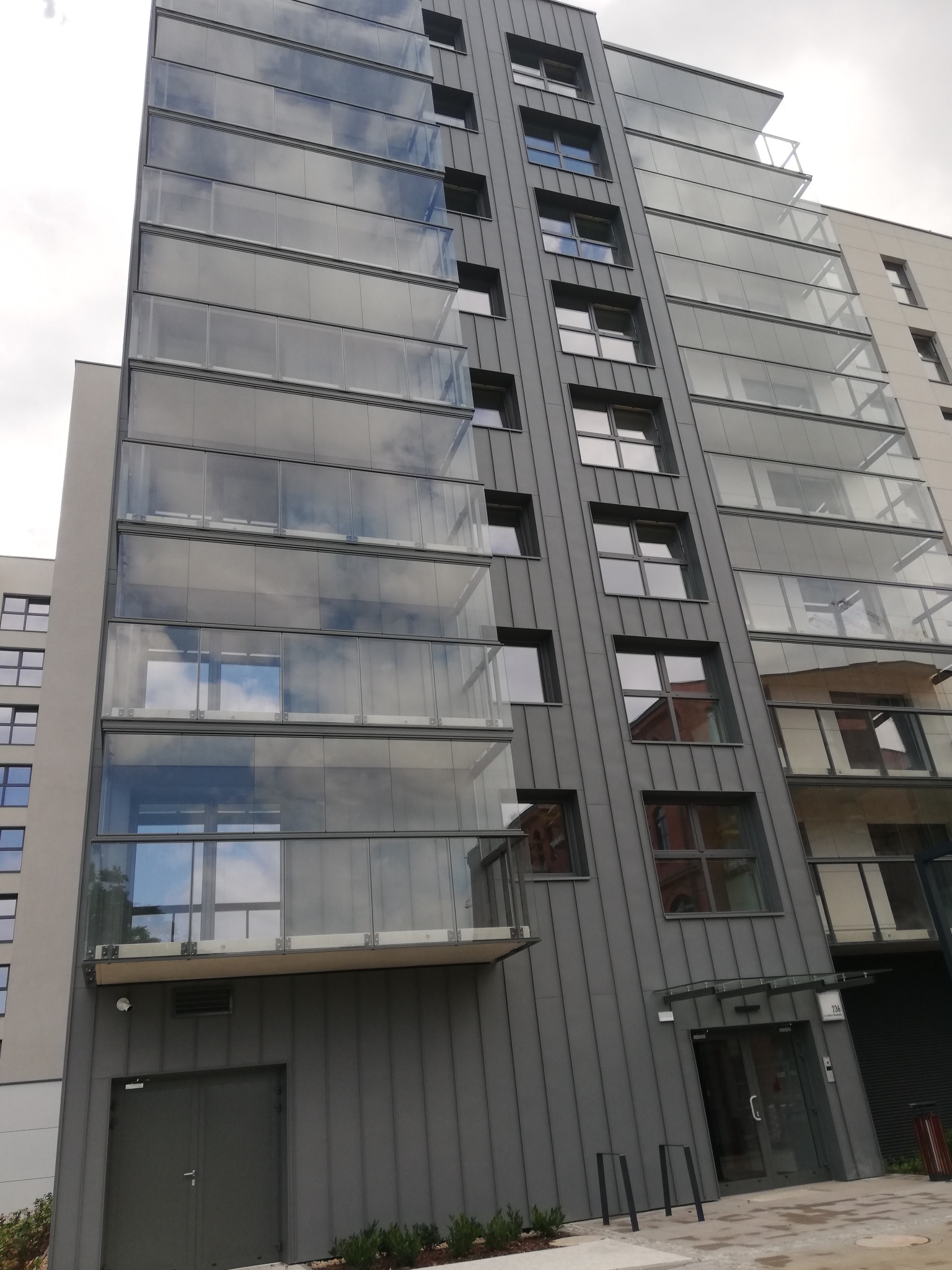 Nowoczesny budynek wielorodzinny z szarymi panelami elewacyjnymi i przeszklonymi balkonami, w których odbija się niebo. Widok z dołu, lekki kąt.