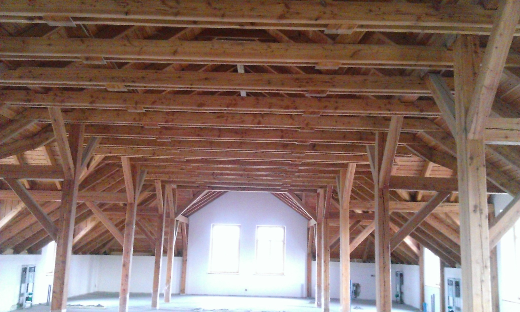Drewniana konstrukcja więźby dachowej z widocznymi belkami i słupami, surowy stan, wnętrze budynku z oknami w tle. Jasne drewno, surowy montaż.