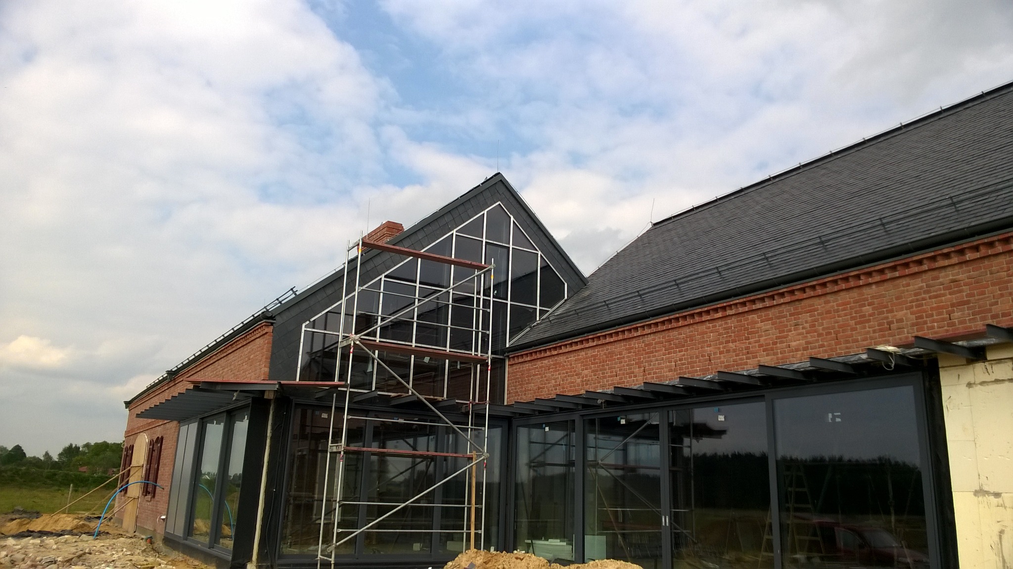 Nowoczesny dom z cegły z ciemną dachówką i dużymi oknami, częściowo zasłonięty rusztowaniem. Widoczne niebo z chmurami i fragment krajobrazu.