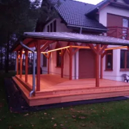 Zadaszony taras z drewna połączony z domem, oświetlony ledami, widok z boku na trawnik i las w tle.