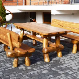 Rustykalny zestaw mebli ogrodowych z litego drewna: stół i dwie ławki z oparciami, na tle brukowanej nawierzchni i budynku z cegły.