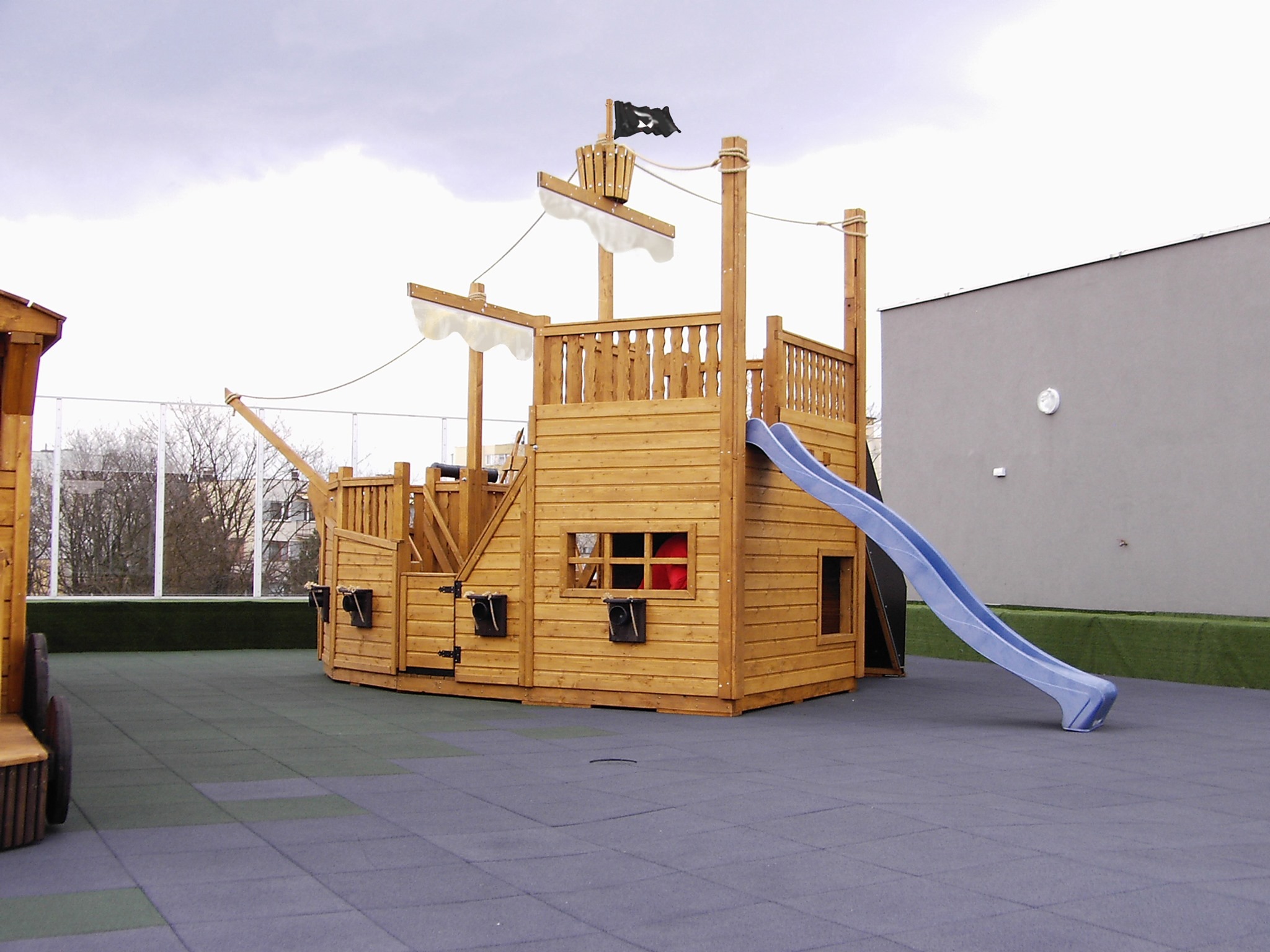 Drewniany plac zabaw w kształcie statku pirackiego z niebieską zjeżdżalnią na bezpiecznej nawierzchni z gumowych płyt.