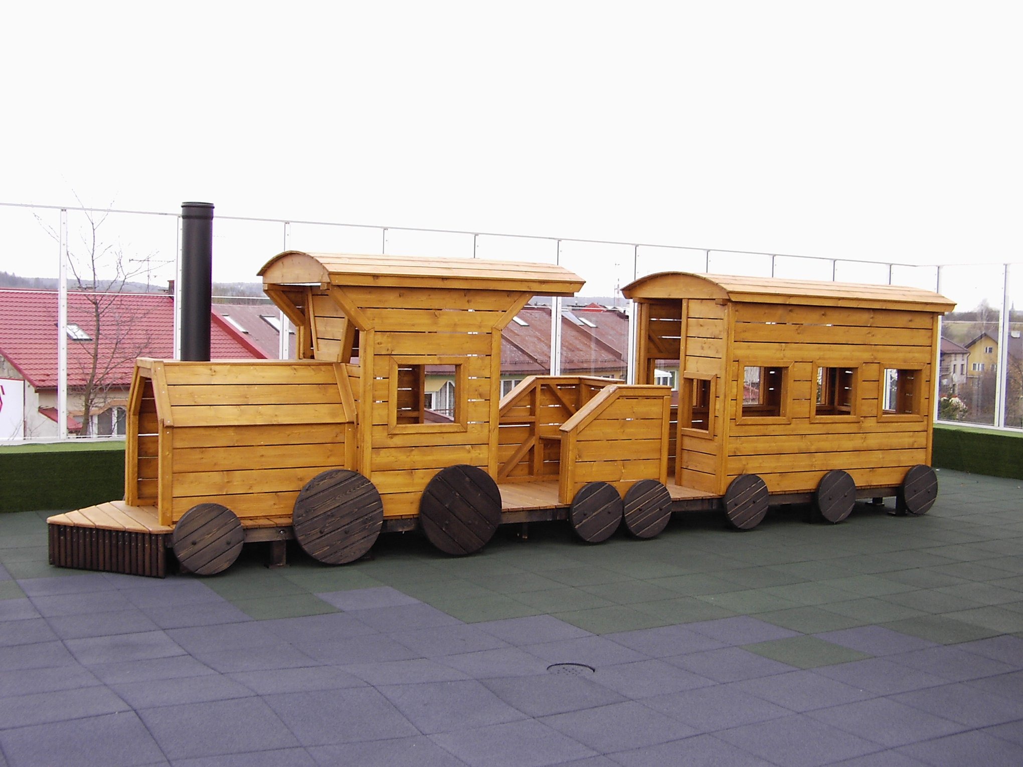 Duża, drewniana lokomotywa z wagonami na placu zabaw z gumową nawierzchnią, z widocznymi detalami konstrukcyjnymi i elementami dekoracyjnymi.