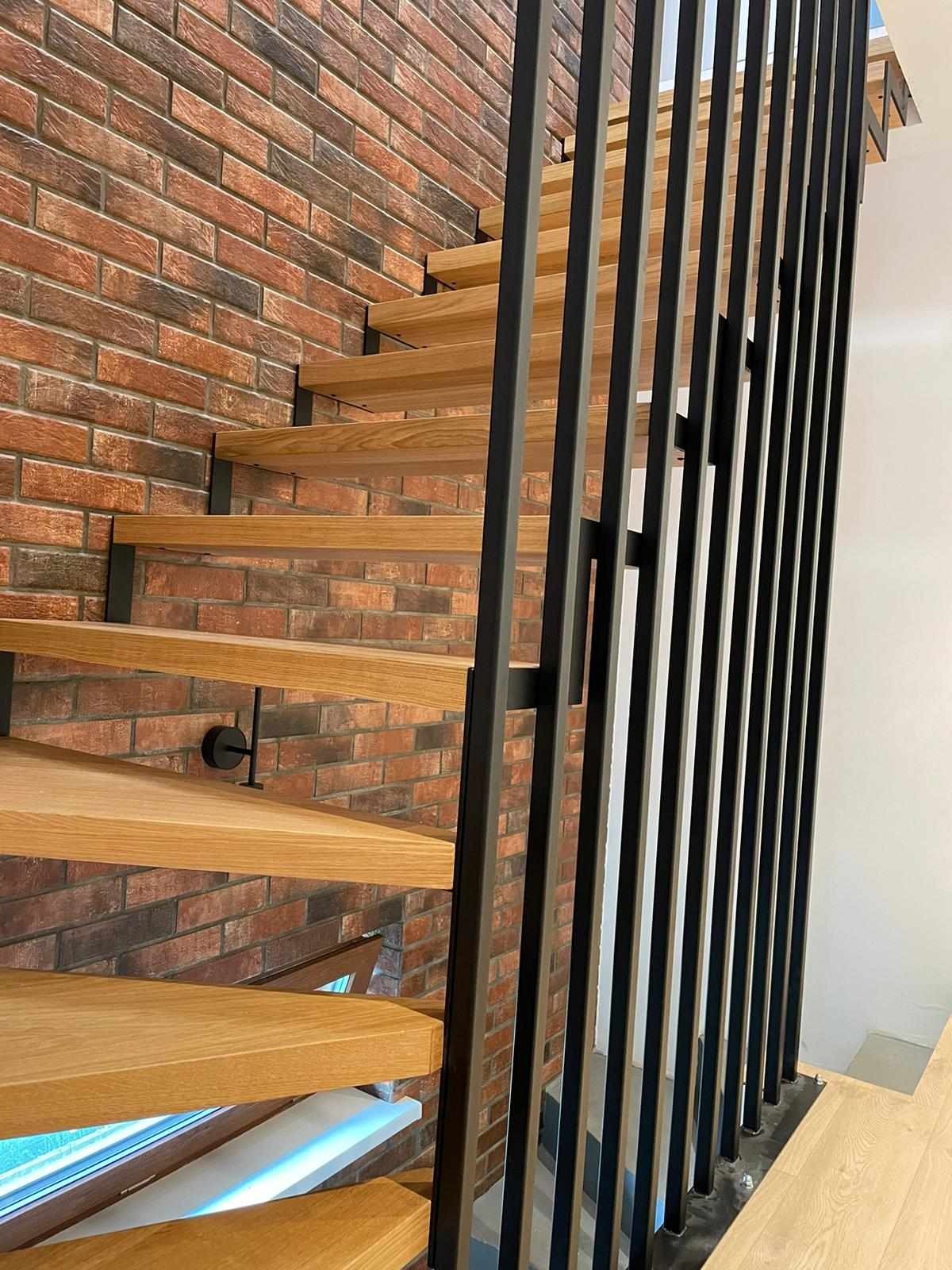 Nowoczesne schody z jasnego drewna z czarną metalową balustradą na tle ściany z cegły klinkierowej.