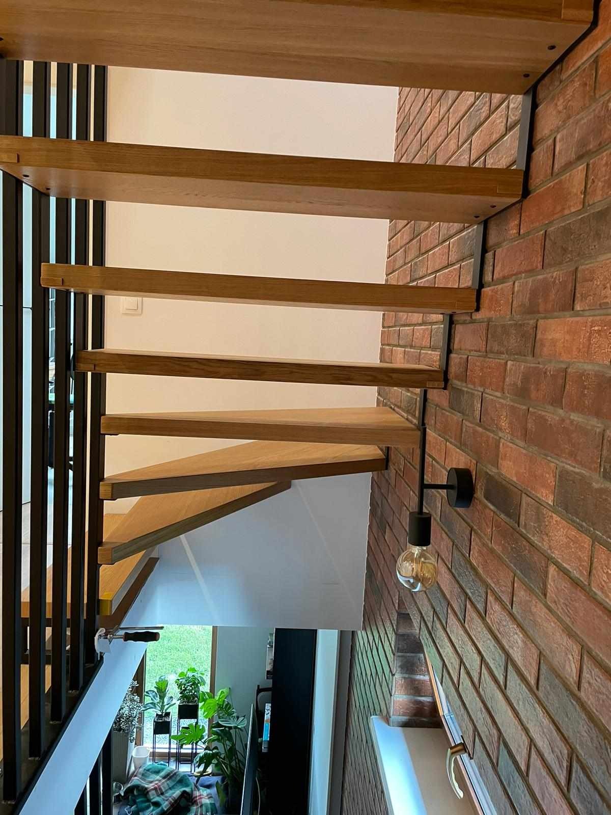 Współczesne schody z dębowymi stopniami, z czarnymi metalowymi wspornikami i balustradą, zamontowane na ceglanej ścianie z designerską lampą edisonowską.