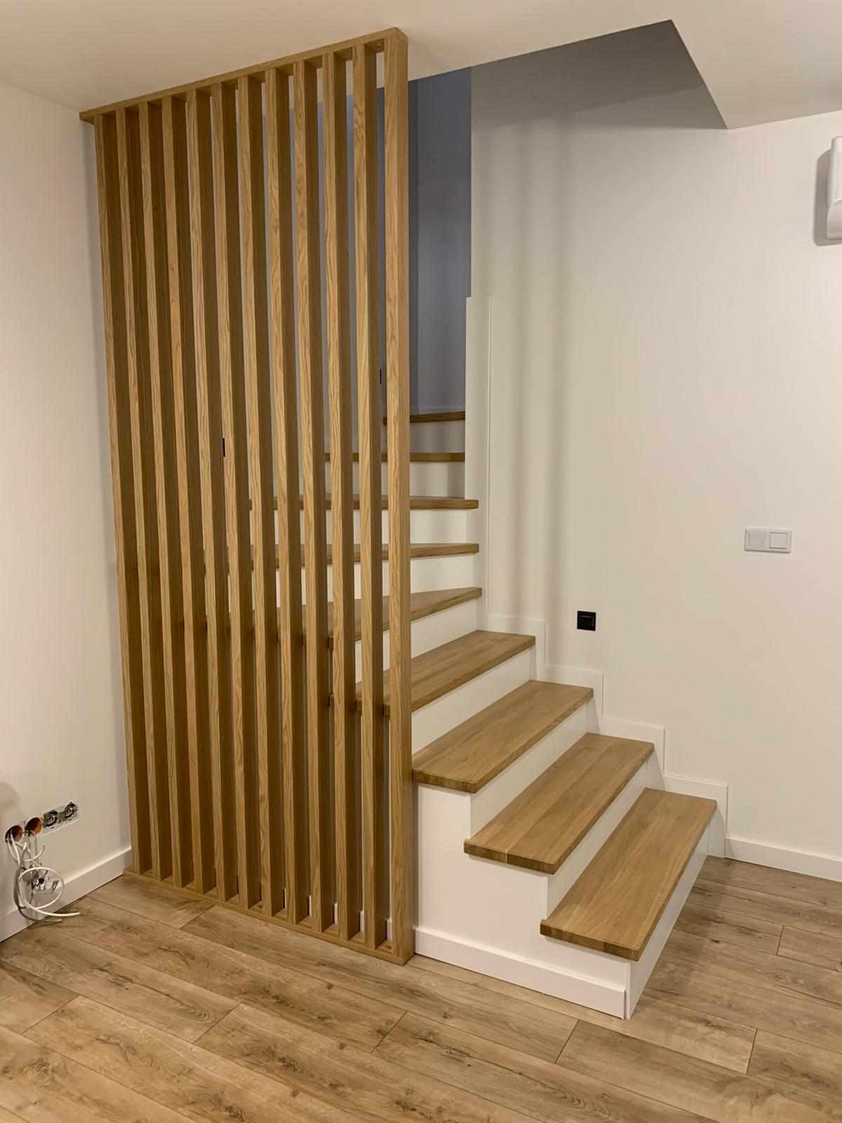 Nowoczesne schody z drewnianymi stopniami i białymi podstopnicami, oddzielone od przestrzeni drewnianą ażurową ścianką działową, widok na drewnianą podłogę.