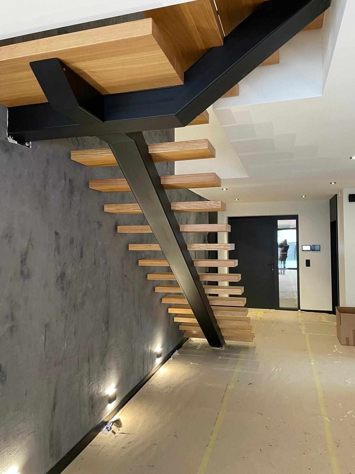 Nowoczesne schody wspornikowe z drewnianymi stopniami i czarną metalową konstrukcją, oświetlenie punktowe wzdłuż ściany, widok na wejście.