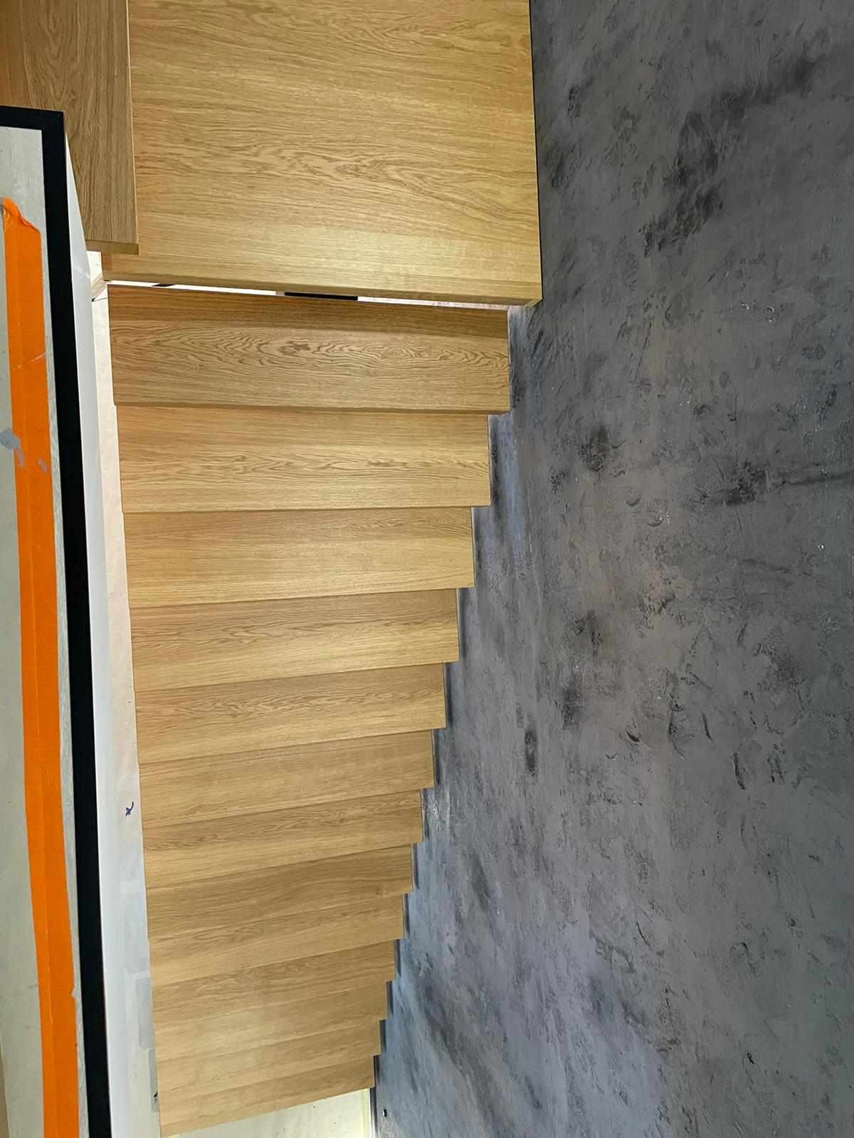 Jasne, drewniane stopnie schodów wewnętrznych, widoczne z boku, kontrastują z szarą, betonową ścianą. Widoczne elementy wykończeniowe, takie jak listwy przypodłogowe i taśma malarska.