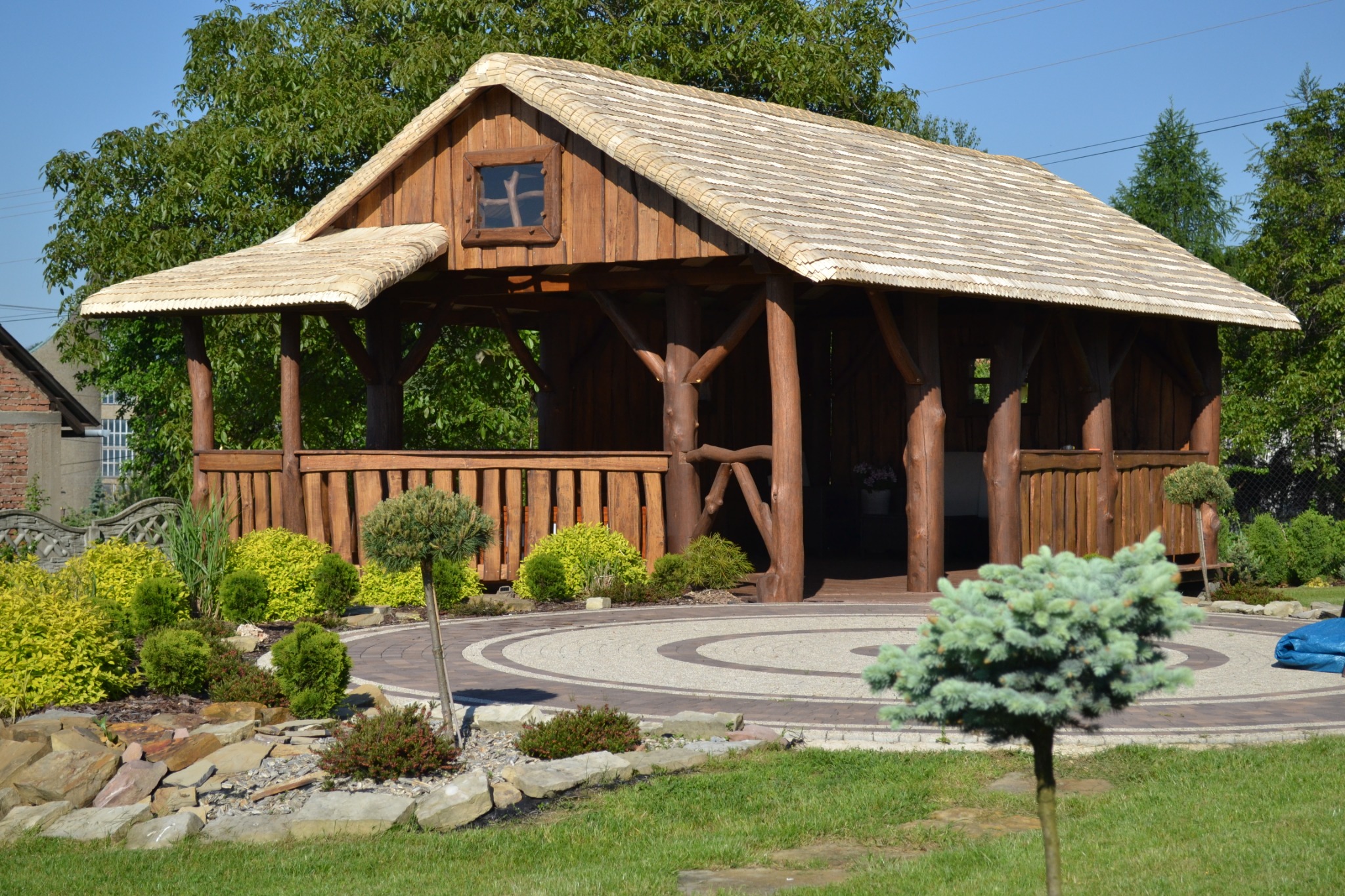 Drewniana altana z krytym dachem, ozdobiona dekoracyjnymi drzewkami i kamieniami w ogrodzie.