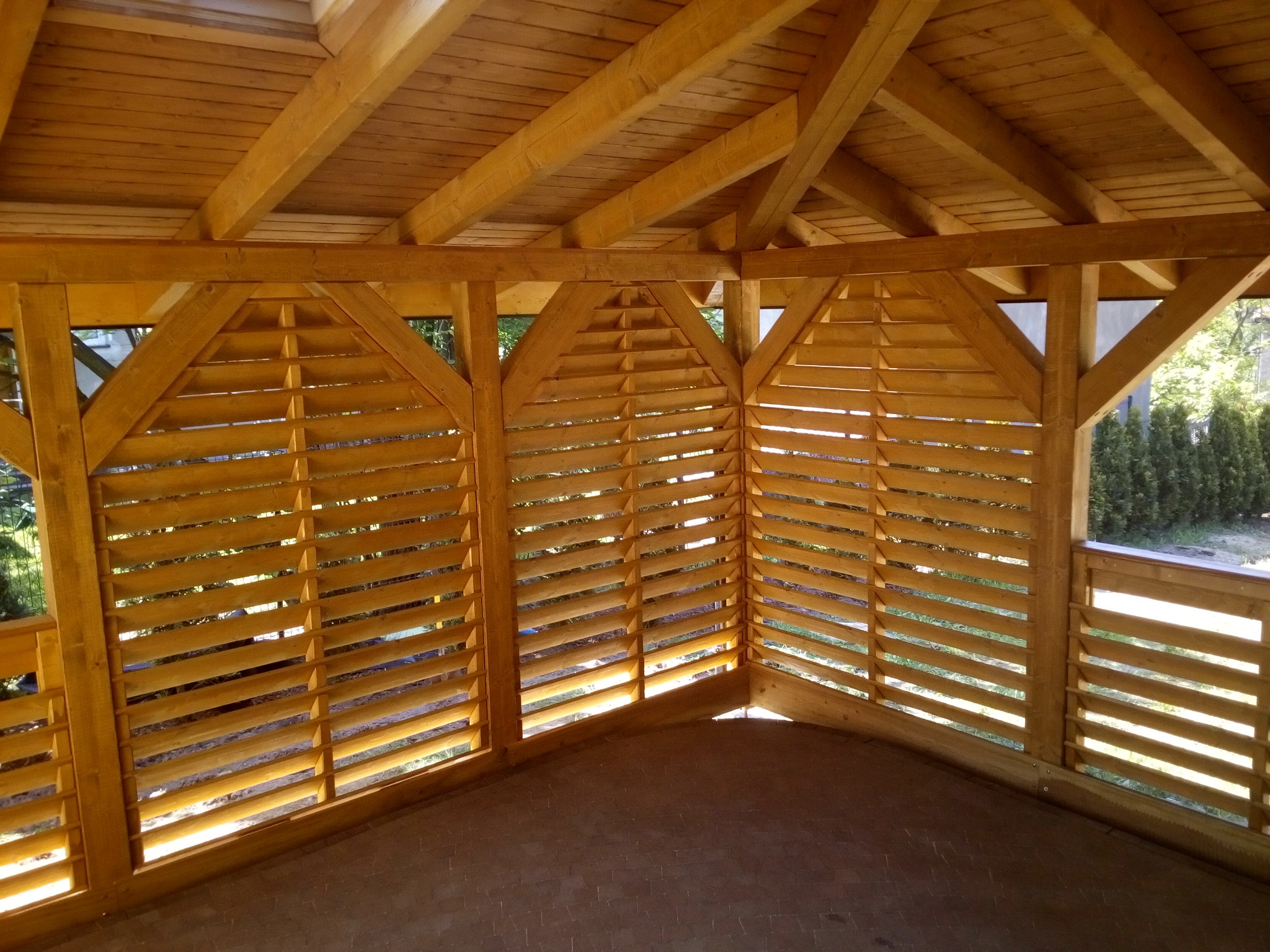Wnętrze drewnianej konstrukcji z ażurowymi ścianami, widoczne elementy dachu i podłoga z kostki brukowej, zieleń w tle.