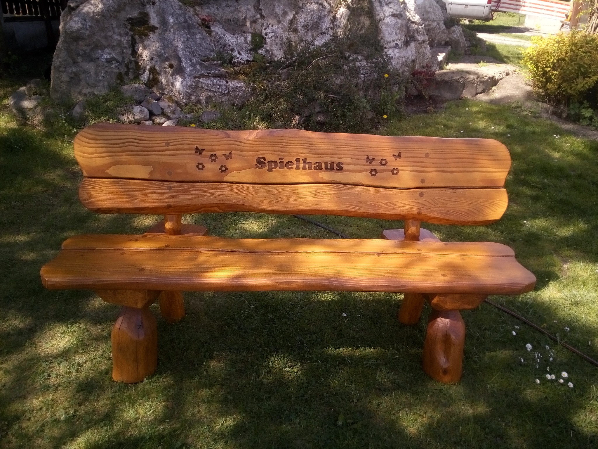 Drewniana ławka ogrodowa z wygrawerowanym napisem 'Spielhaus' i motywami kwiatowymi, stojąca na trawniku przed skalnym ogrodem.