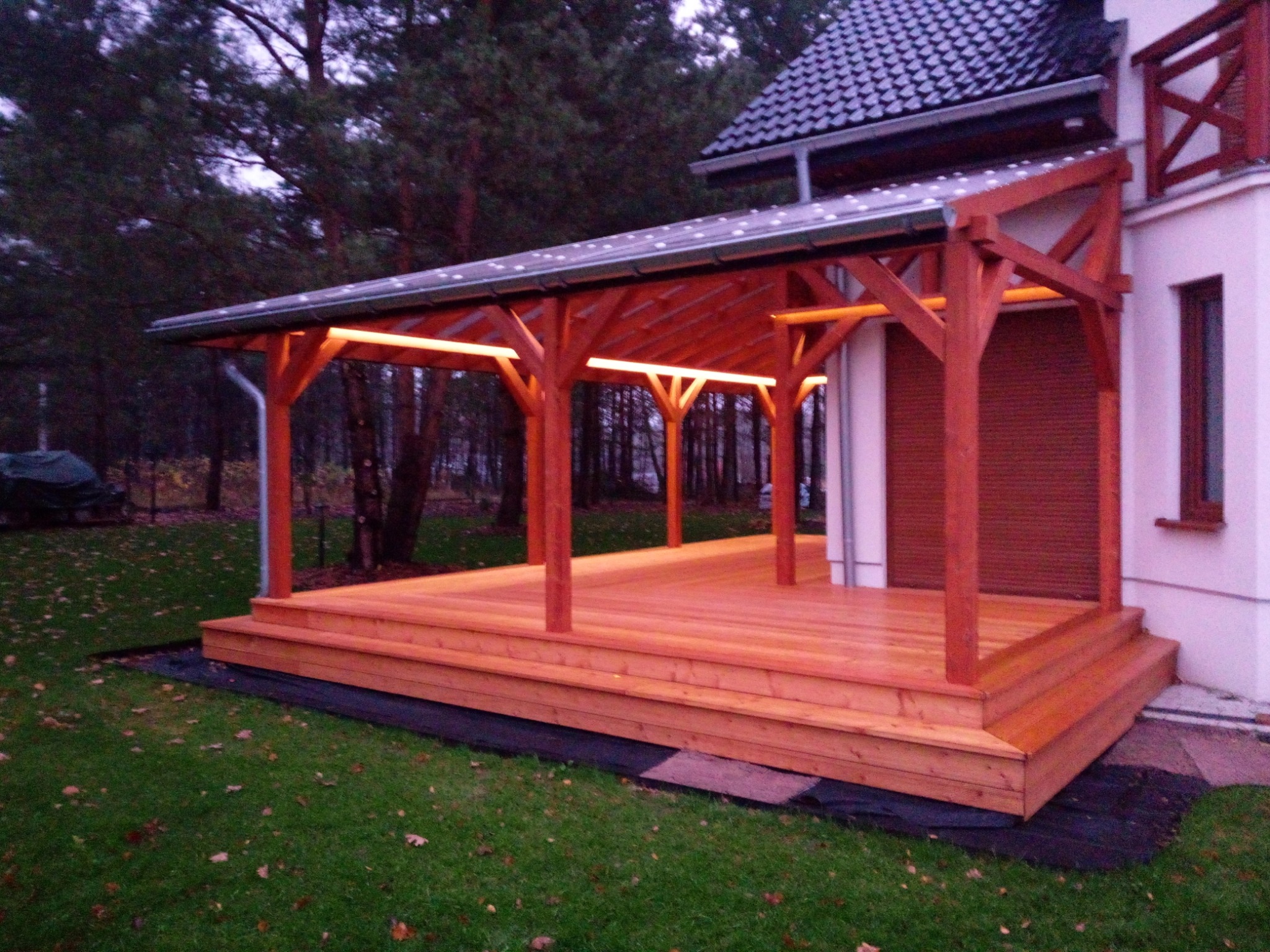 Zadaszony taras z drewna z oświetleniem LED, przylegający do białego domu, otoczony trawnikiem i drzewami iglastymi.