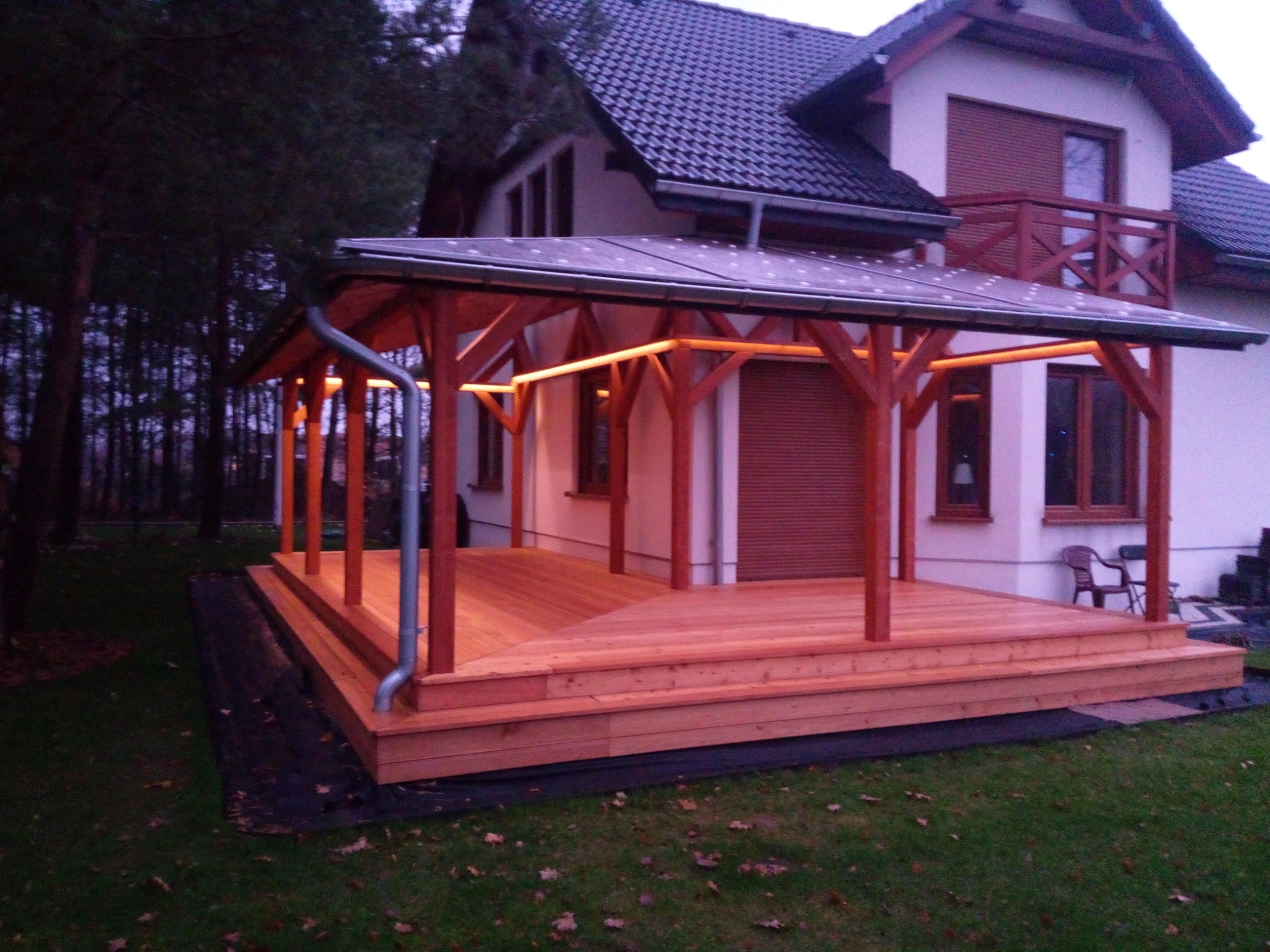 Zadaszony taras z drewna połączony z domem, oświetlony ledami, widok z boku na trawnik i las w tle.