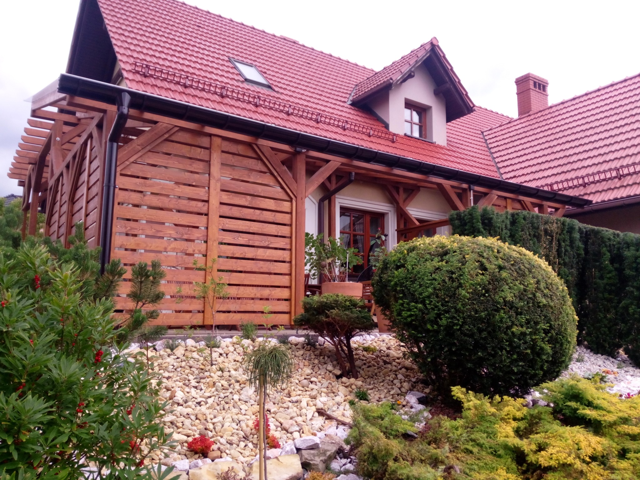 Drewniana konstrukcja osłaniająca taras przylegająca do domu z czerwoną dachówką, otoczona kamieniami i różnorodną roślinnością.