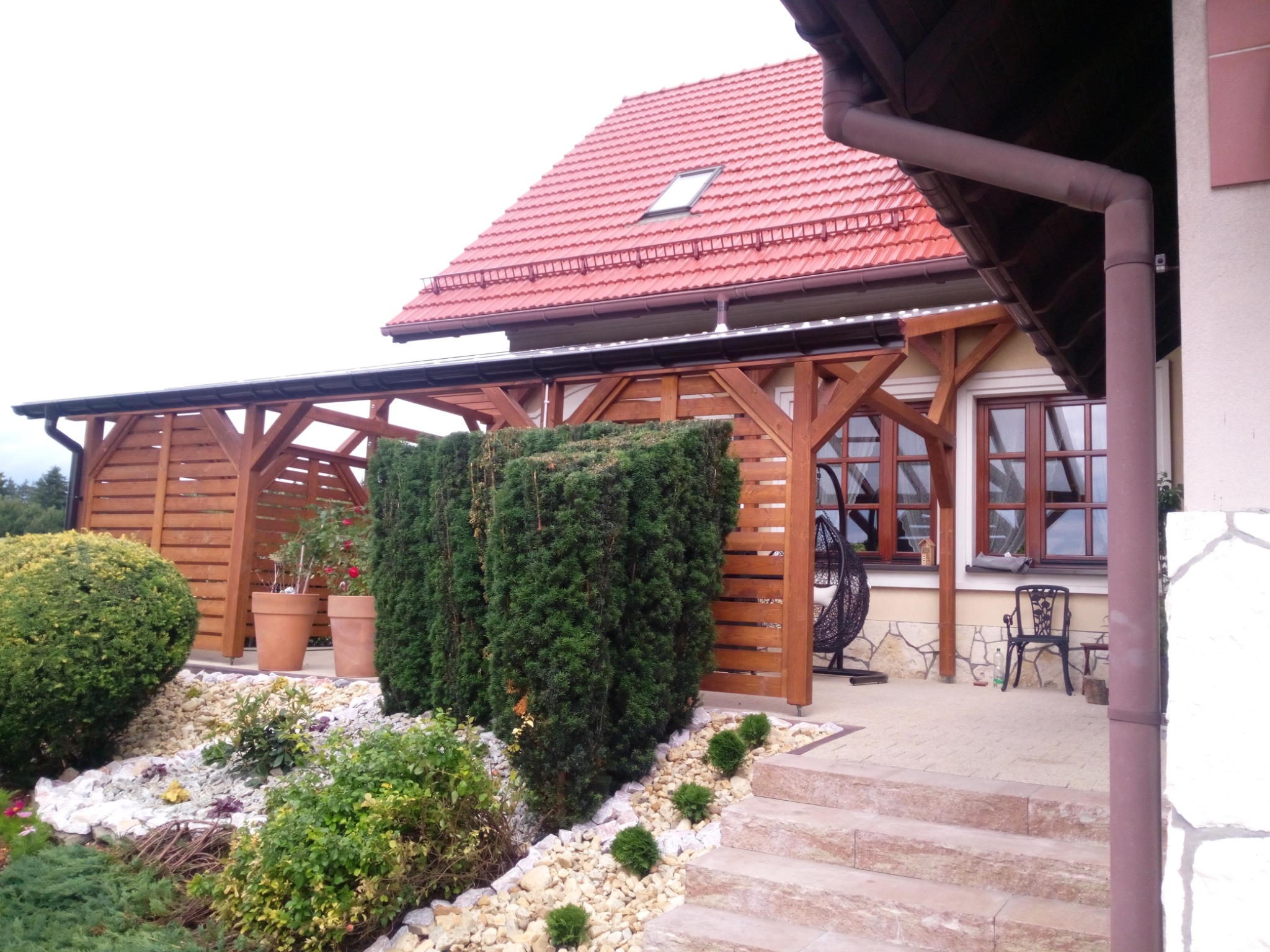 Drewniana pergola z zadaszeniem przylegająca do domu z czerwonym dachem, widoczne donice z kwiatami, ozdobne krzewy i ścieżka z kamieni.
