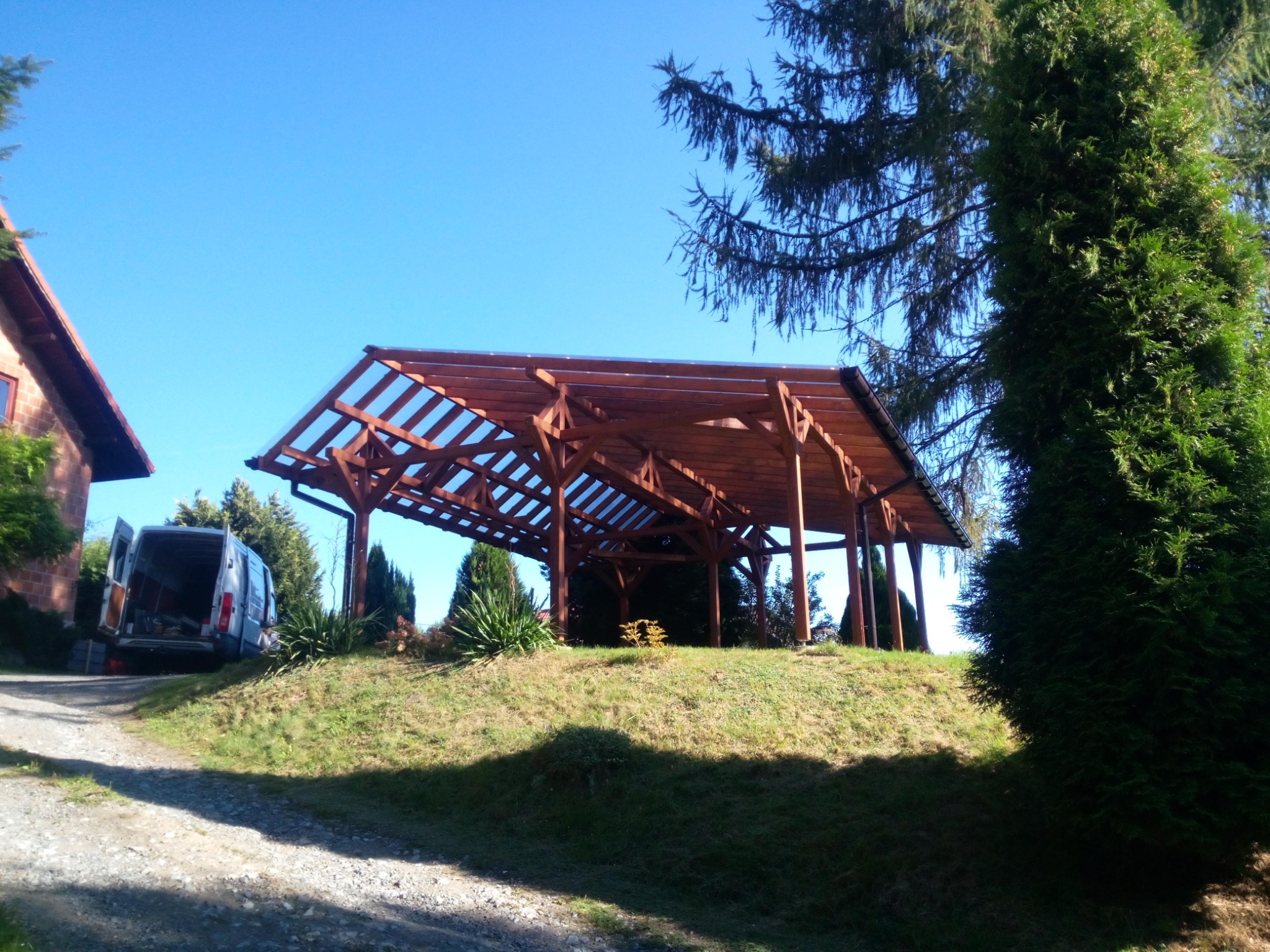 Drewniana konstrukcja z przezroczystym zadaszeniem na wzgórzu, widoczny samochód dostawczy z otwartymi drzwiami w tle, drzewa iglaste po bokach, błękitne niebo.