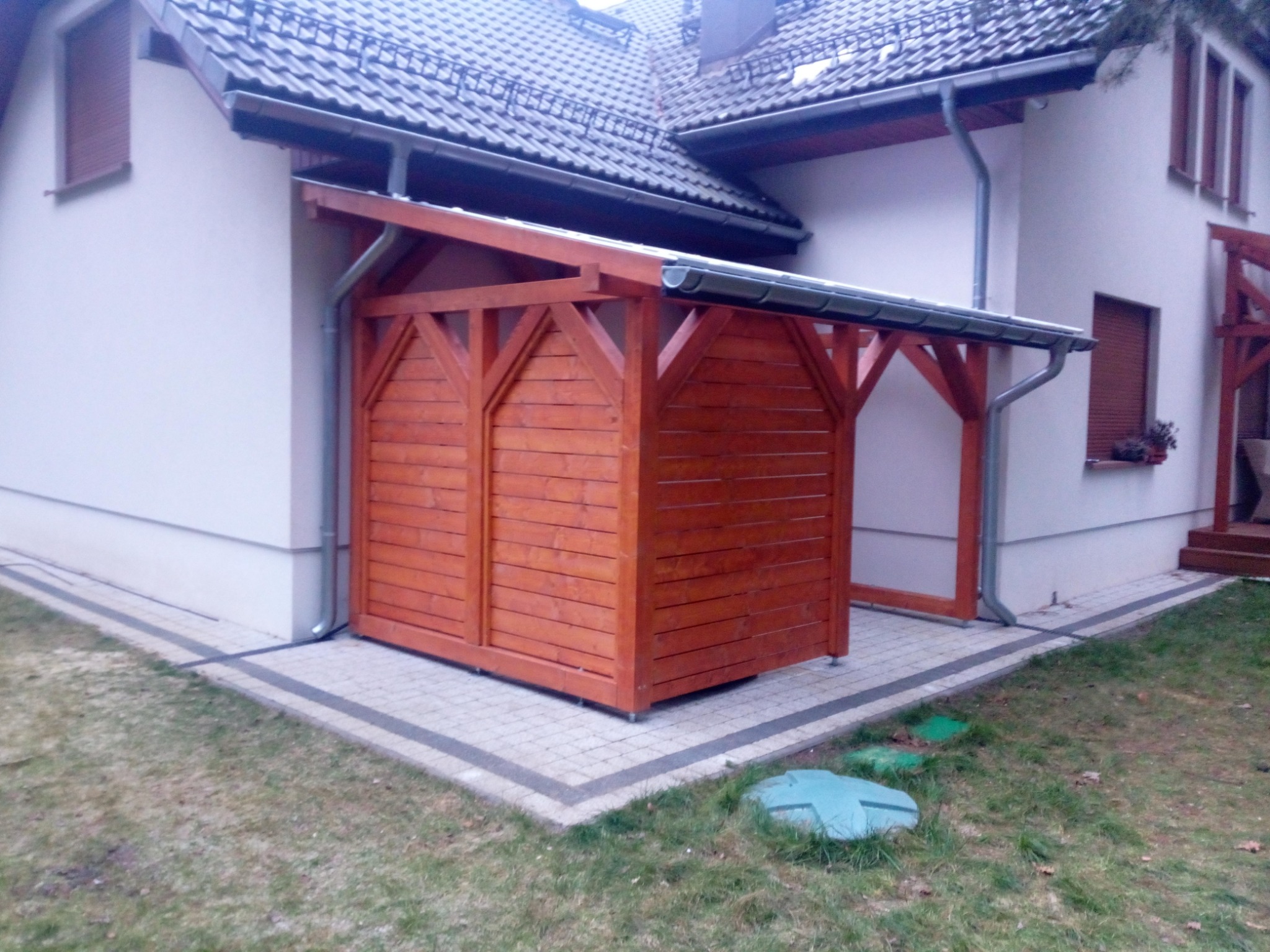 Zabudowana, drewniana konstrukcja z zadaszeniem przylegająca do ściany domu, z widocznym orynnowaniem, na kostce brukowej, otoczona trawnikiem.