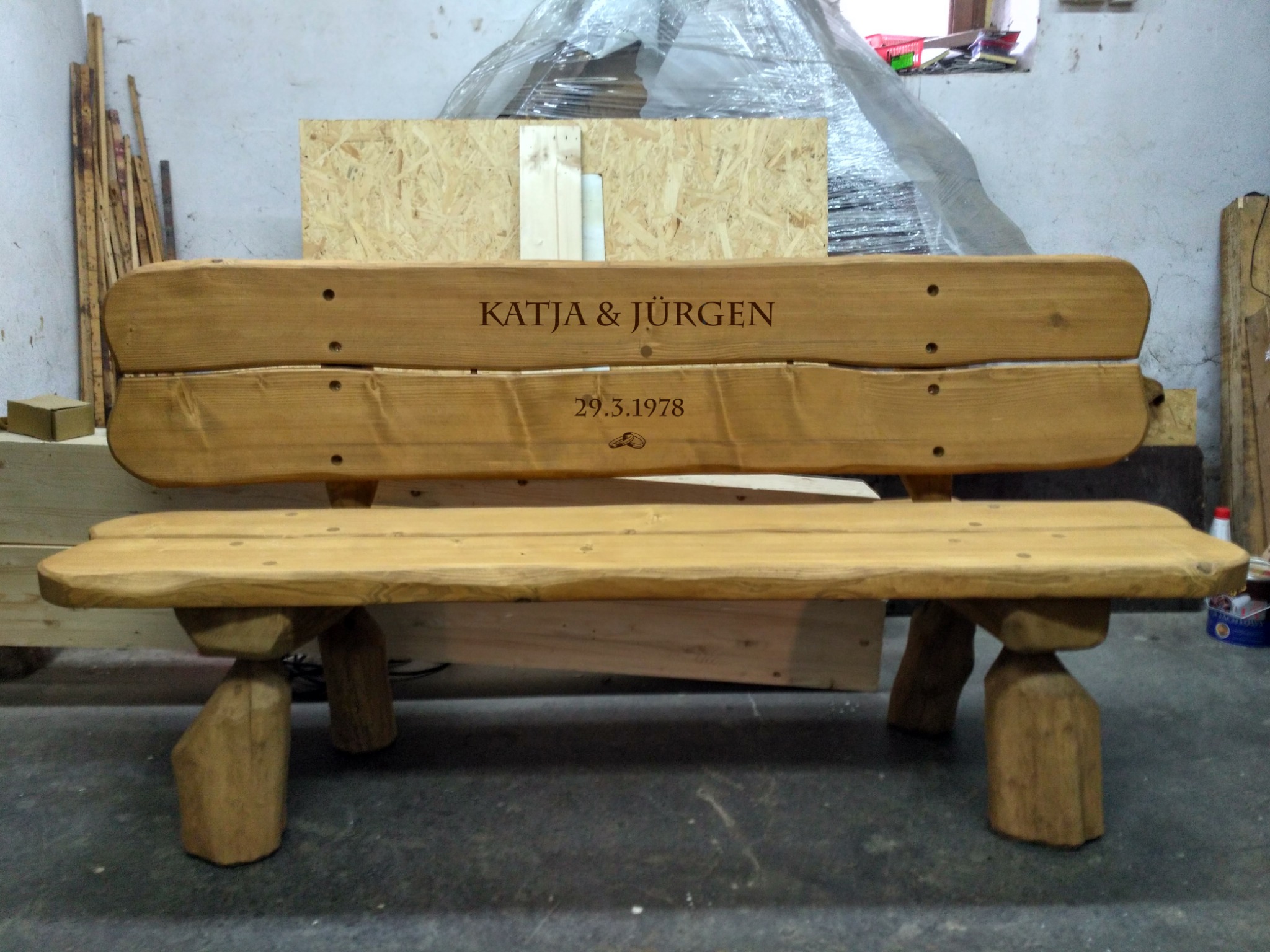 Rustykalna ławka z jasnego drewna z wygrawerowanym napisem 'Katja & Jürgen' i datą 29.3.1978, stojąca w warsztacie.