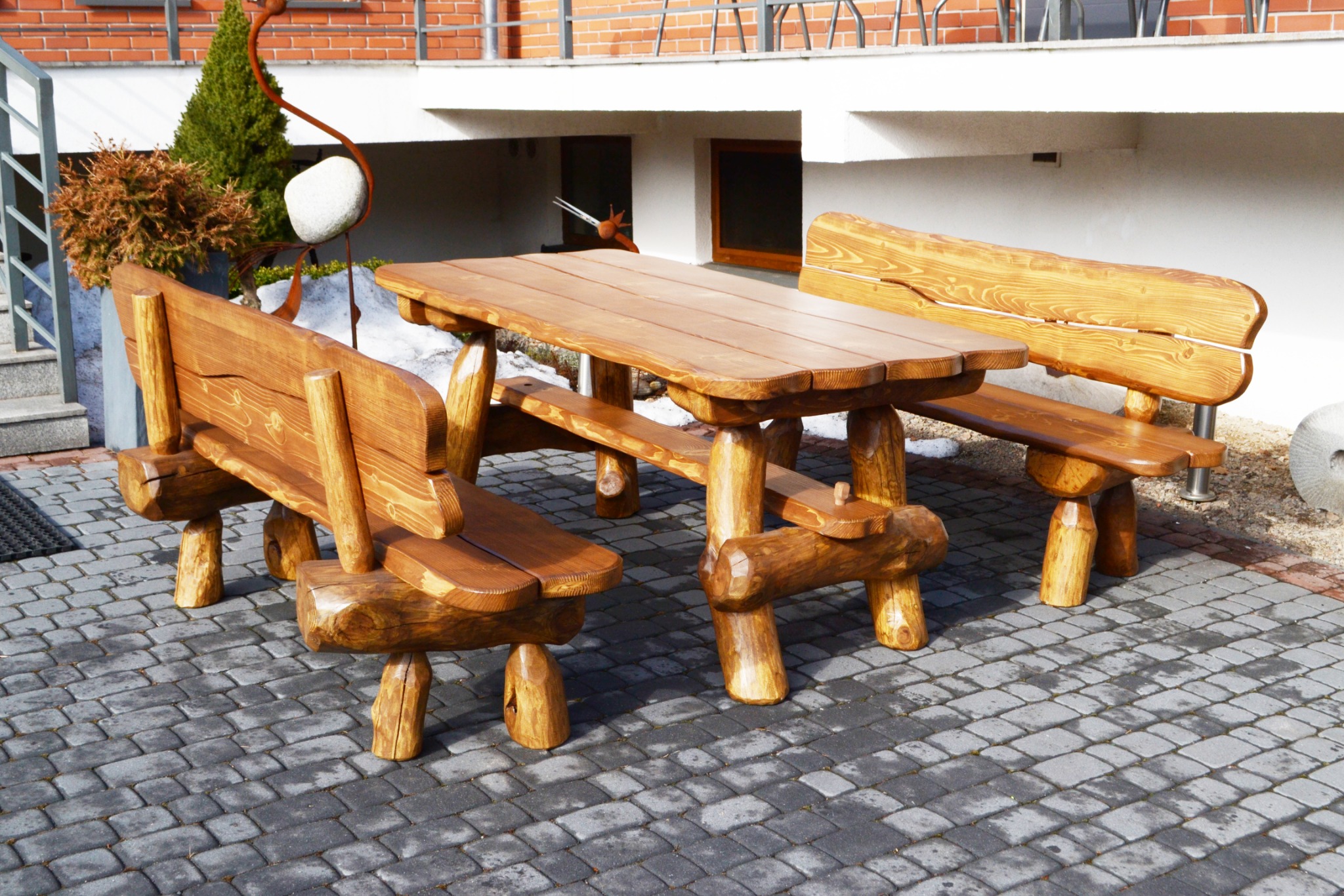 Rustykalny zestaw mebli ogrodowych z litego drewna: stół i dwie ławki z oparciami, na tle brukowanej nawierzchni i budynku z cegły.