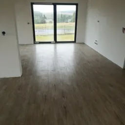 Nowo położone panele podłogowe w jasnym odcieniu drewna w pustym pokoju z widokiem na zewnątrz przez duże okno balkonowe.