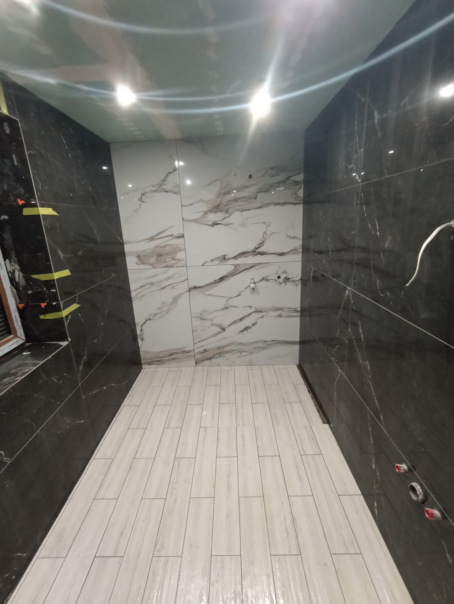 Wnętrze łazienki w trakcie prac wykończeniowych, widoczne czarne i białe płytki imitujące marmur na ścianach oraz jasne płytki imitujące drewno na podłodze, fragment okna z żółtą taśmą...