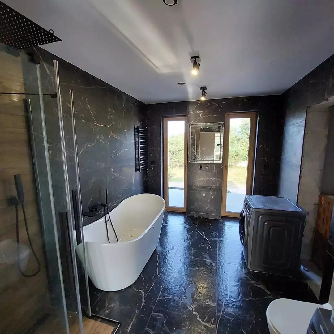 Nowoczesna łazienka wykończona czarnym marmurem, z wolnostojącą białą wanną, kabiną prysznicową typu walk-in z czarną armaturą, pralką oraz grzejnikiem drabinkowym.