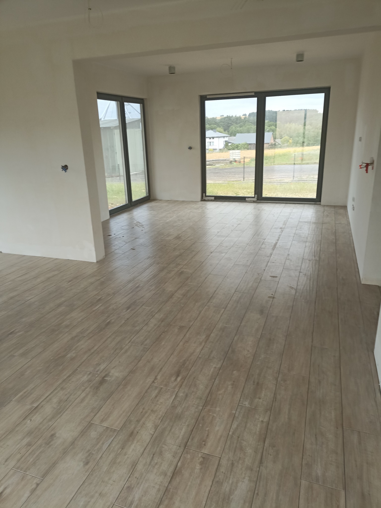 Jasne, podłużne płytki imitujące drewno ułożone w dużym, pustym pomieszczeniu z widokiem na zewnątrz przez duże okna przesuwne.