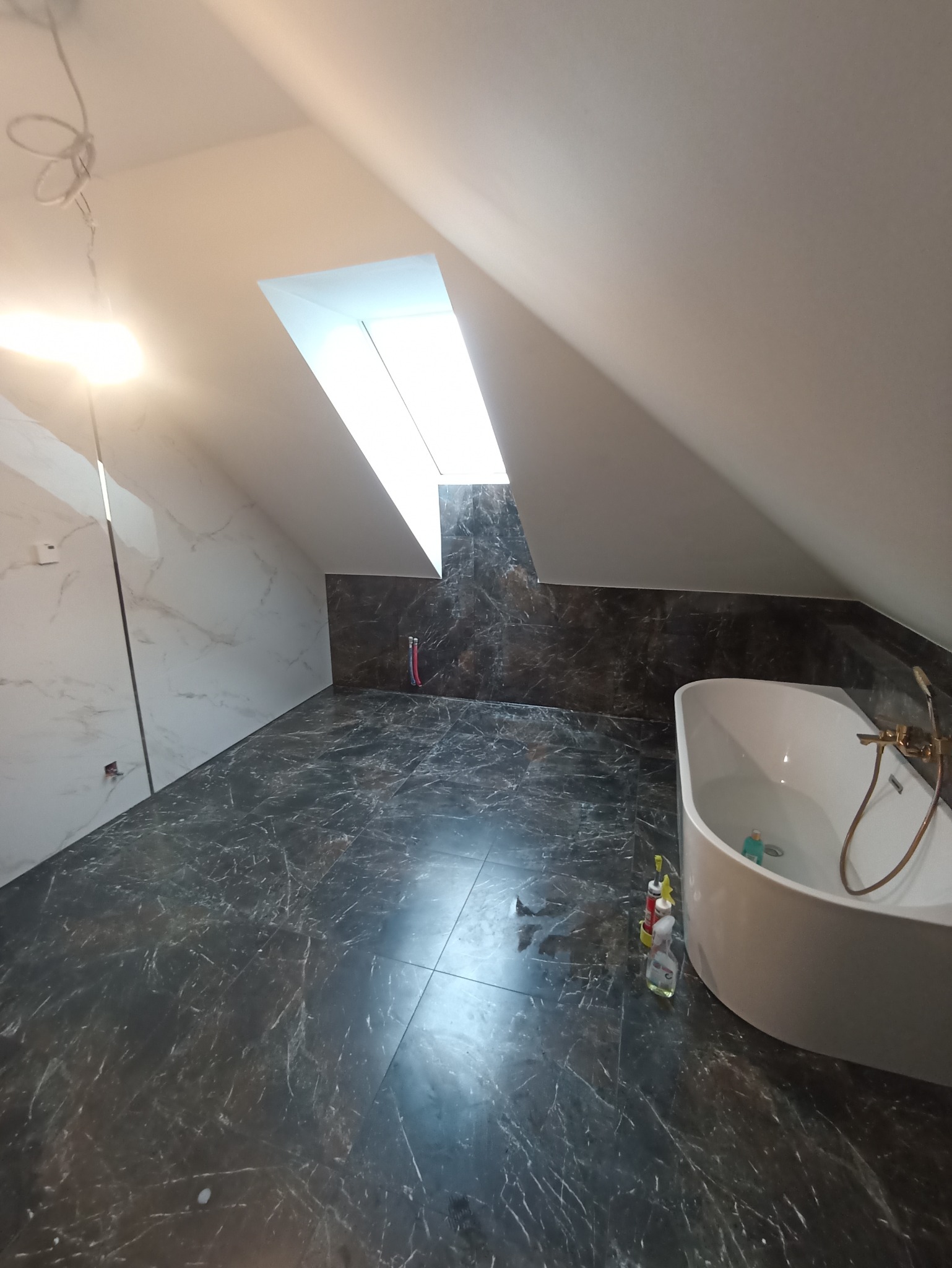 Ukończona łazienka na poddaszu z marmurowymi płytkami w odcieniach szarości i bieli, owalną wanną z złotą armaturą, oświetlona światłem wpadającym przez okno dachowe, widoczne środki czystości...