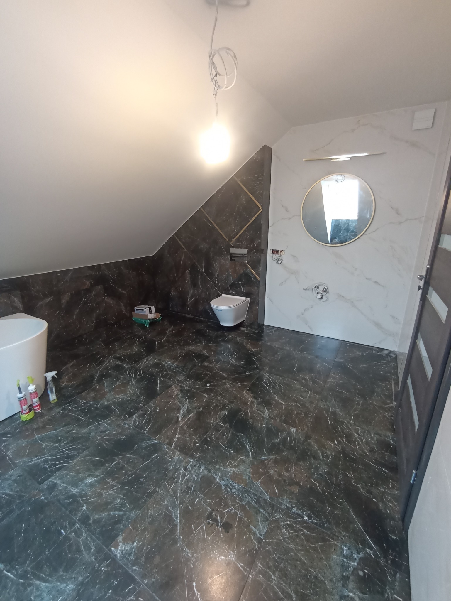 Ukończona łazienka na poddaszu z marmurowymi płytkami w odcieniach szarości i bieli, widoczna toaleta, okrągłe lustro z oświetleniem, biała wanna i narzędzia na podłodze.