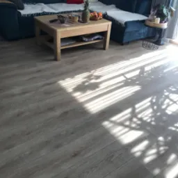 Ułożone panele podłogowe w salonie z widocznym fragmentem sofy, stolika kawowego i padającego światła słonecznego tworzącego cienie na podłodze.