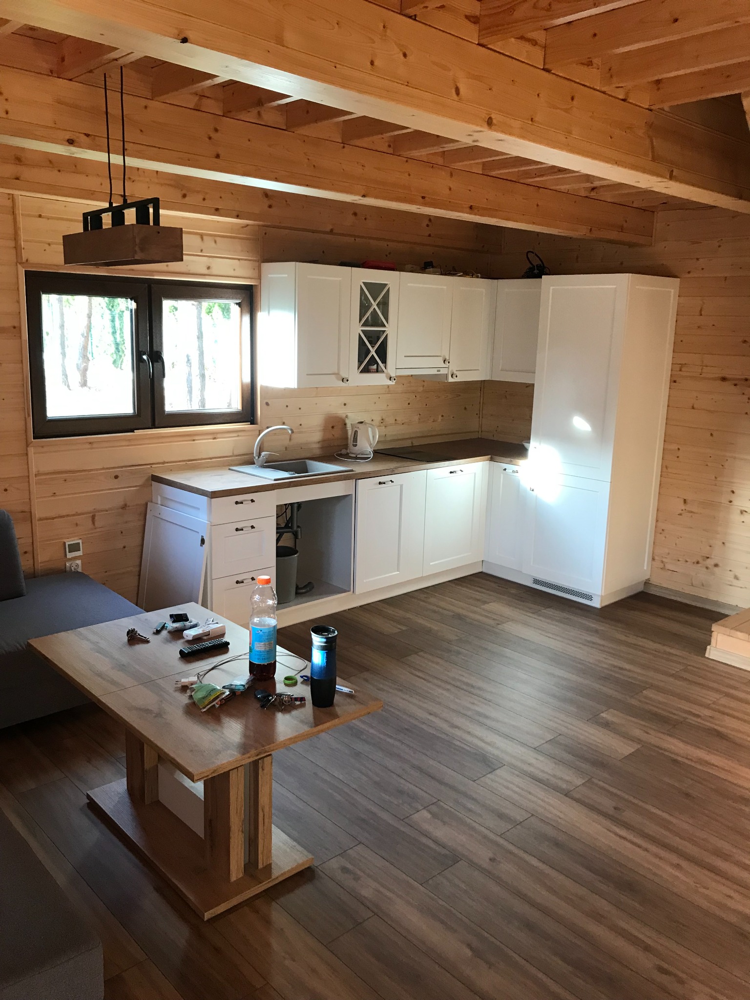 Drewniany domek z wykończoną kuchnią: białe szafki, drewniane ściany i podłoga imitująca panele, stół z drobiazgami na pierwszym planie.