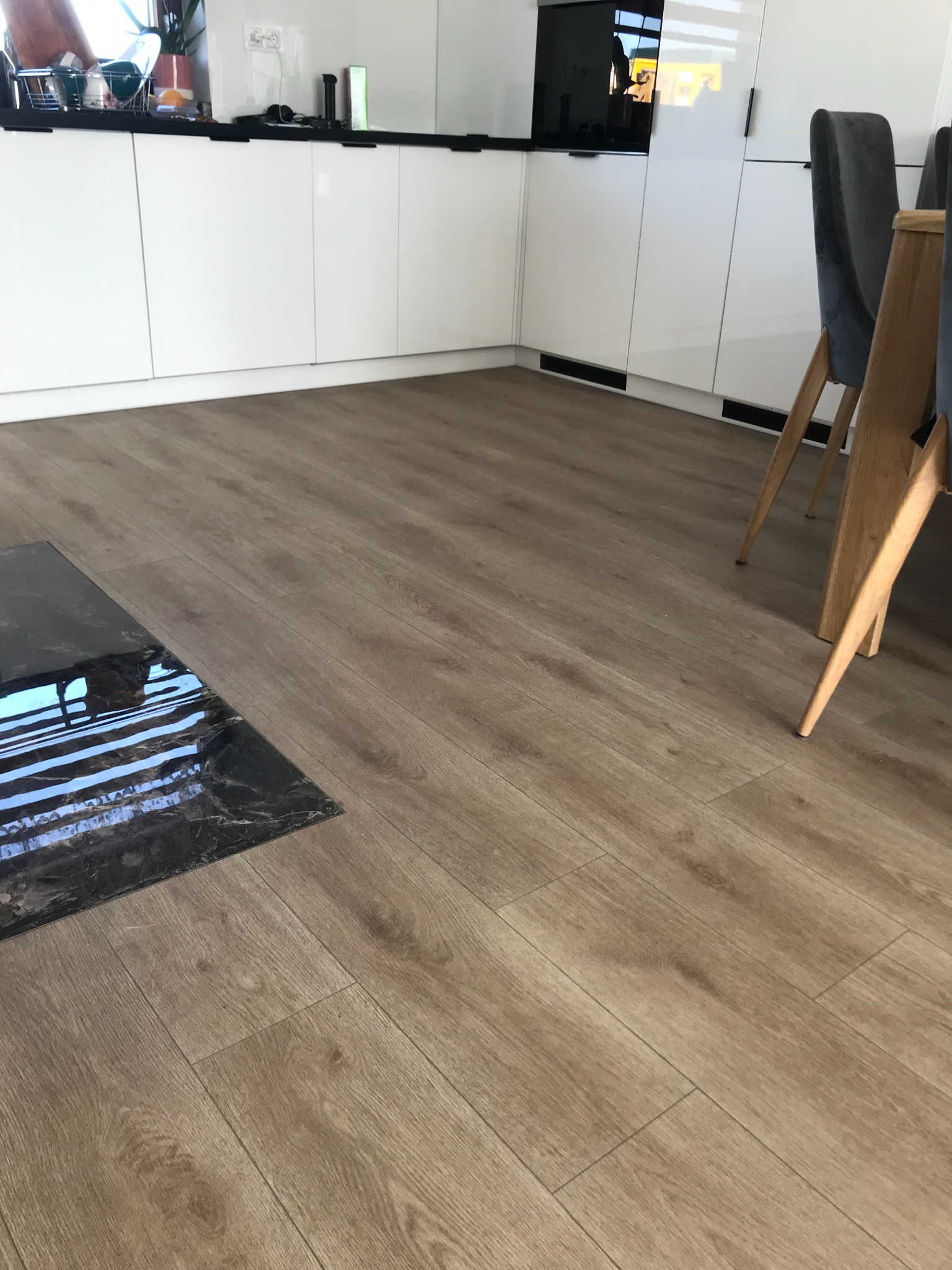 Jasnobrązowe panele podłogowe w nowoczesnej kuchni z białymi szafkami i czarnym blatem stołu, widoczne krzesła z szarą tapicerką.